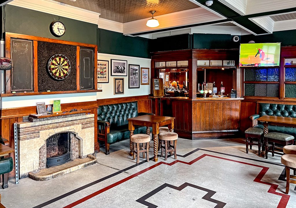 Nursery Inn, Stockport: The vault (public bar) with original tiled fireplace with dart board above, wooden bar counter in the corner and lino floor.