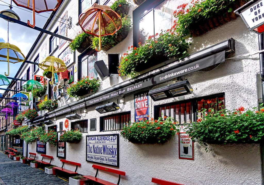 Duke of York, Belfast: North side with red benches, tandem with riders and old signs on wall and umbrellas across the alleyway