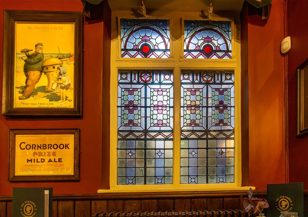 Victoria Hotel, Leeds: Stained glass window with geometric designs next to two small brewery posters.