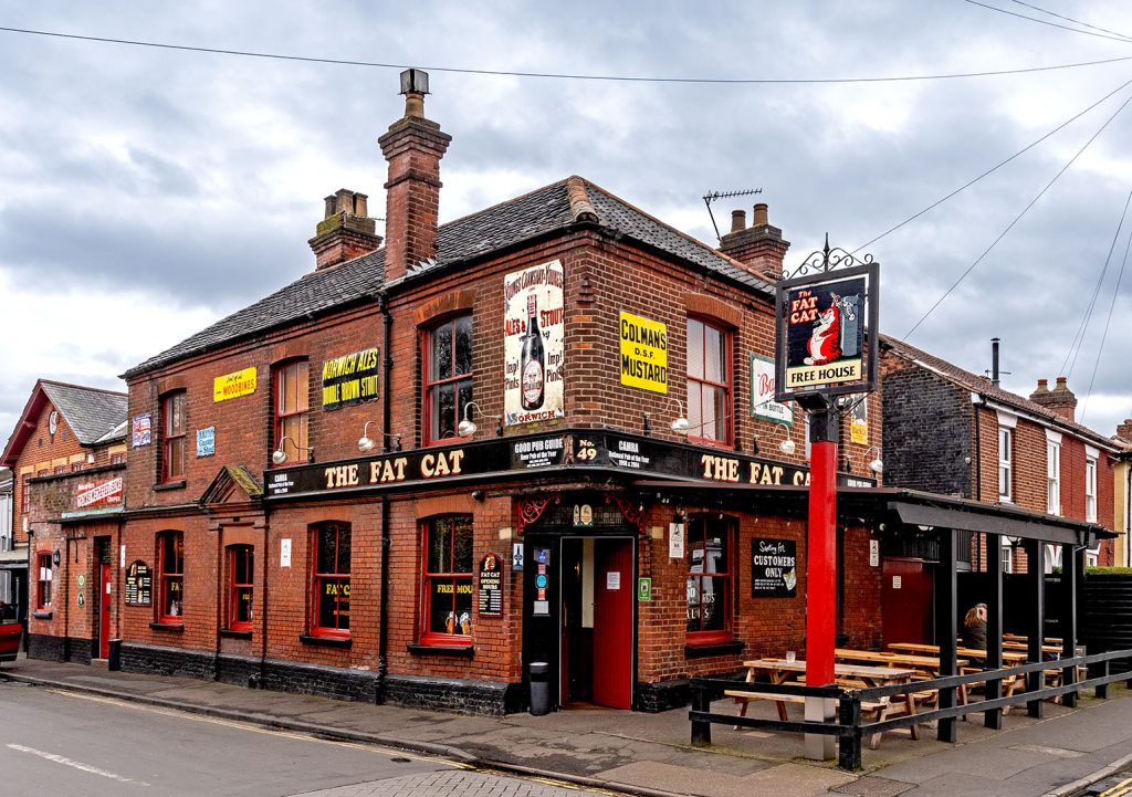 Fat Cat, Norwich: Full exterior of red brick corner pub with vintage metal advertising signs on walls.
